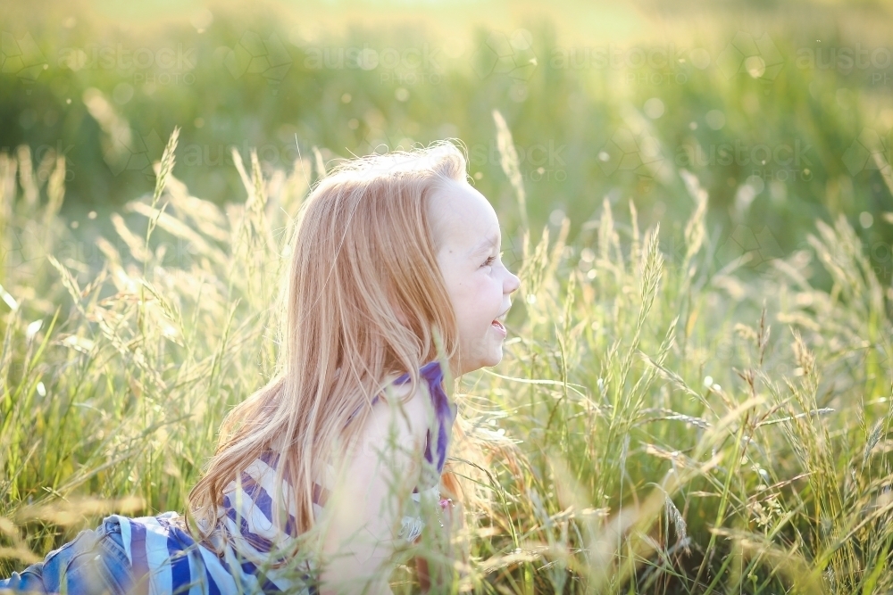 Profile view of young girl lying in long grass with big smile in golden afternoon sunlight - Australian Stock Image