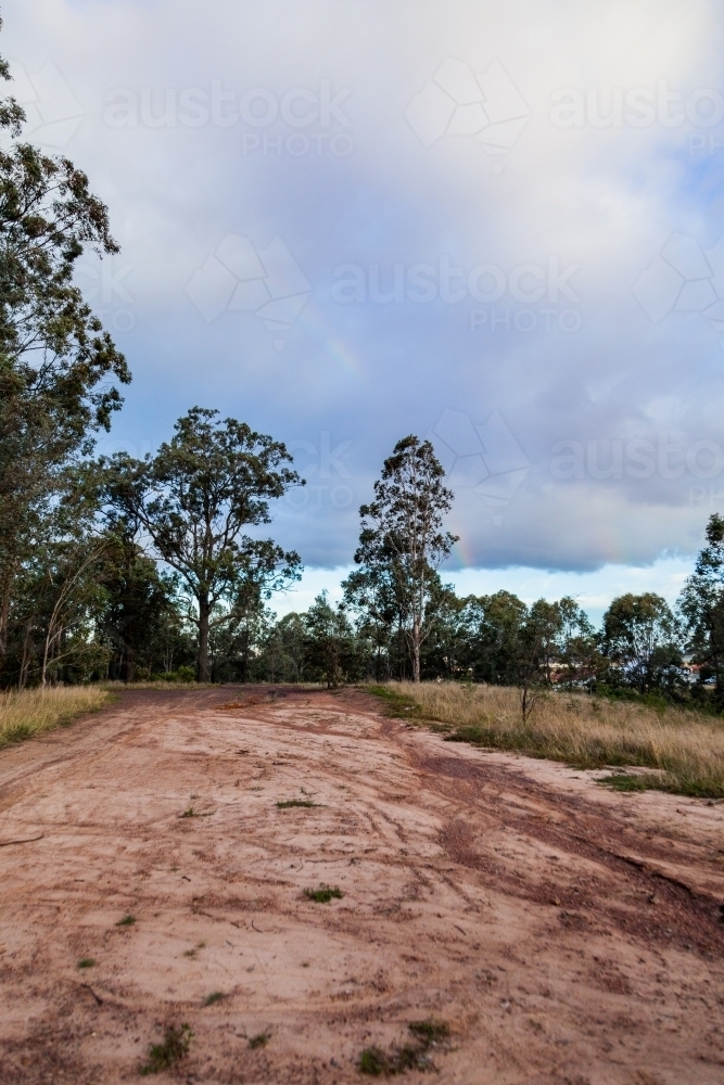 rainbow in rain clouds above rural dirt road scene in paddock - Australian Stock Image