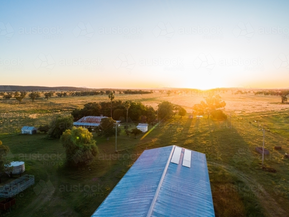 Rays of setting sun touching solar panels on farm shed roof, trees casting shadows over homestead - Australian Stock Image