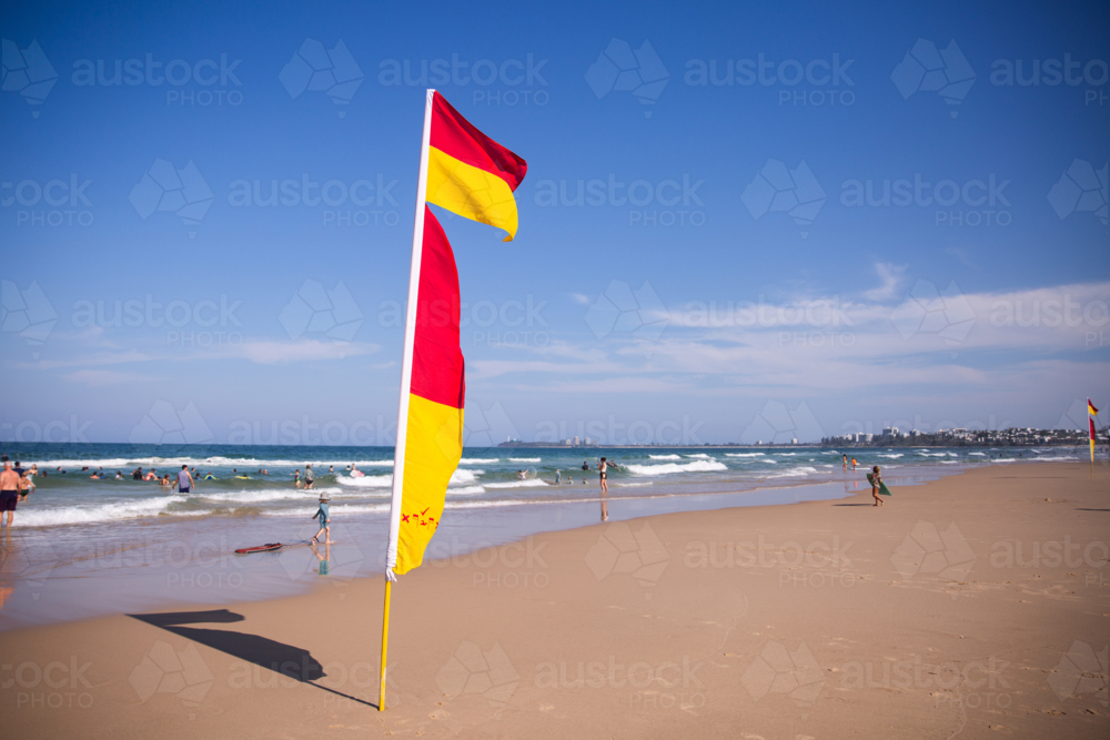 Red and yellow beach safety flags on a sunny summer beach scene with people wading in water - Australian Stock Image