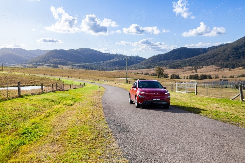 Red electric vehicle car driving down rural country road on sunlit day - Australian Stock Image