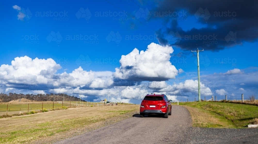 Red electric vehicle car driving down rural country road on sunlit day - Australian Stock Image