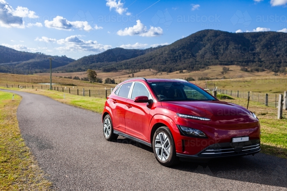 Red electric vehicle car driving down rural country road on sunlit day - Australian Stock Image