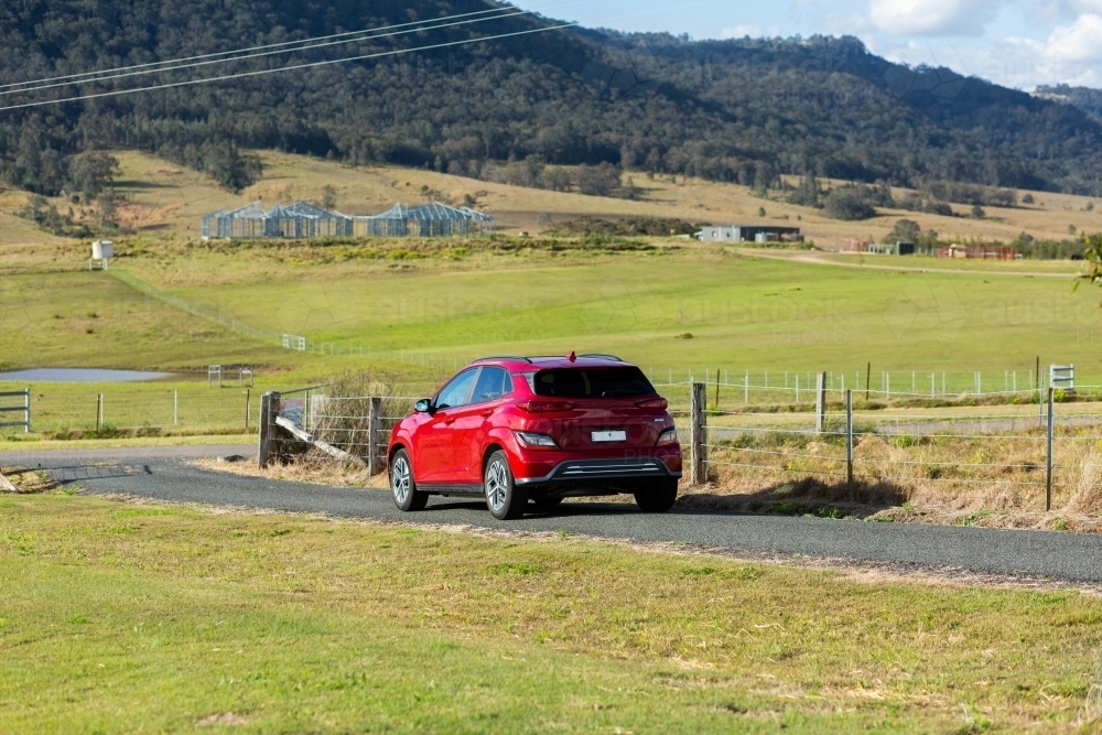 Red electric vehicle car driving down rural country road on sunlit day - Australian Stock Image