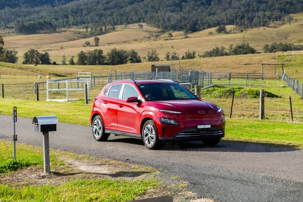 Red electric vehicle car driving down rural country road on sunlit day - Australian Stock Image