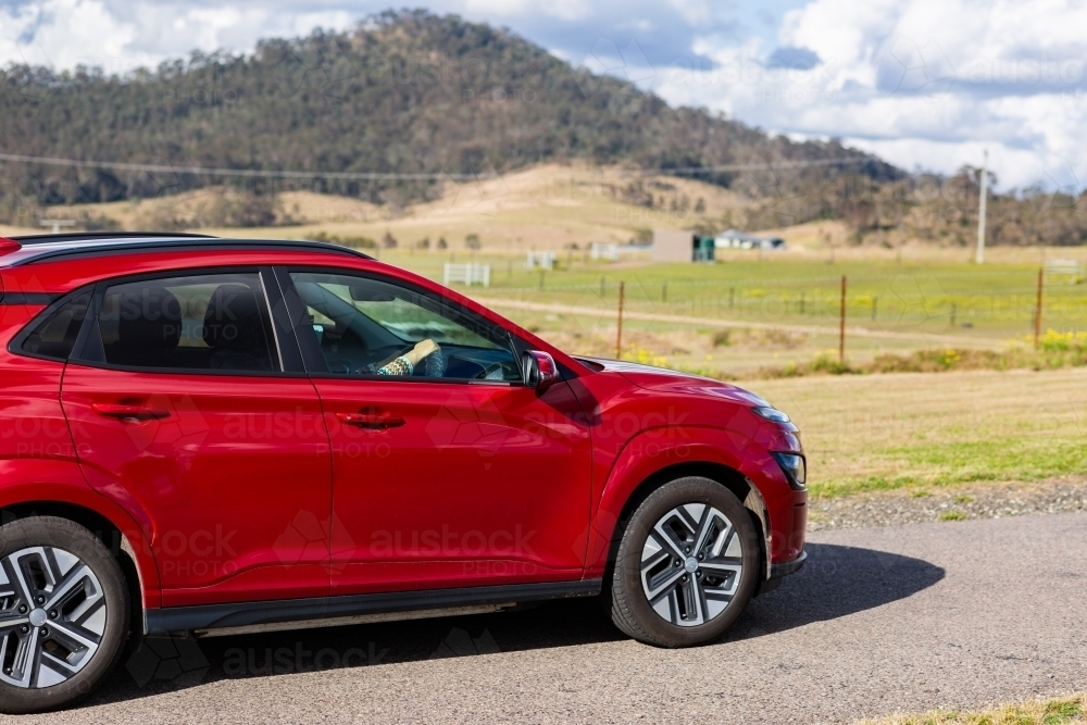 Red electric vehicle car driving down rural country road on sunlit day - Australian Stock Image