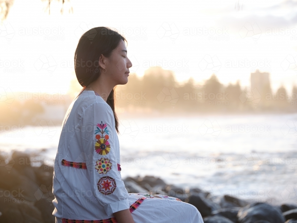 Relaxed Chinese woman sitting on rocks by the shore in the evening - Australian Stock Image