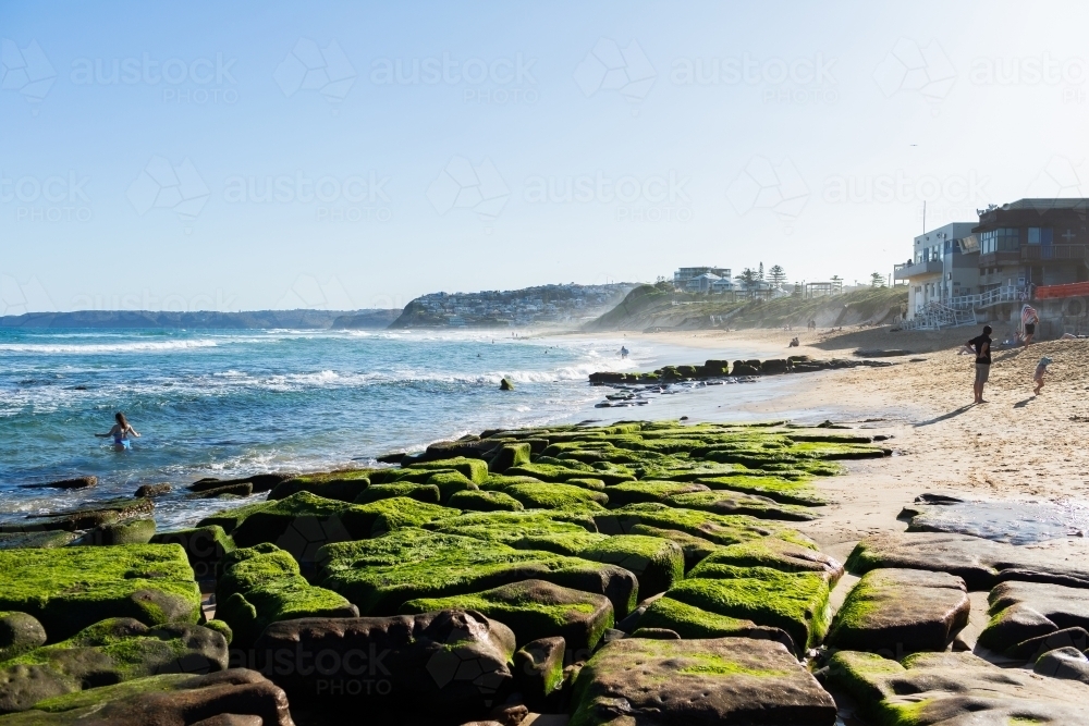 rocks on Bar Beach with sea spray in the air on spring afternoon with beachgoers in water - Australian Stock Image