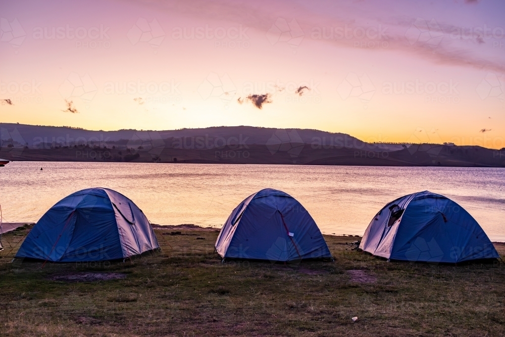 Row of tents at lakeside campground with dusk sky reflecting in lake water - Australian Stock Image