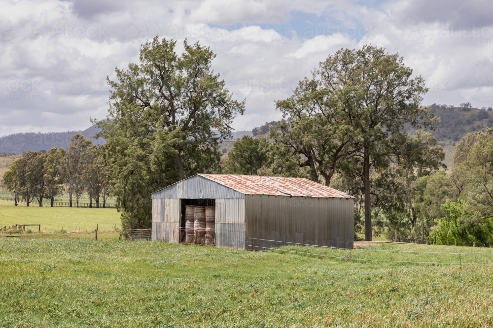 Rural scene with hay bales stacked in an old barn with trees & hills in the background - Australian Stock Image