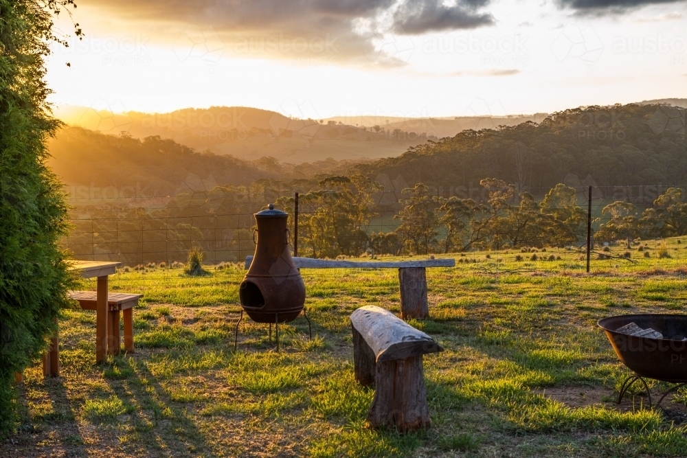 Rural setting at sunset with fireplace and seating overlooking a valley and mountain range - Australian Stock Image