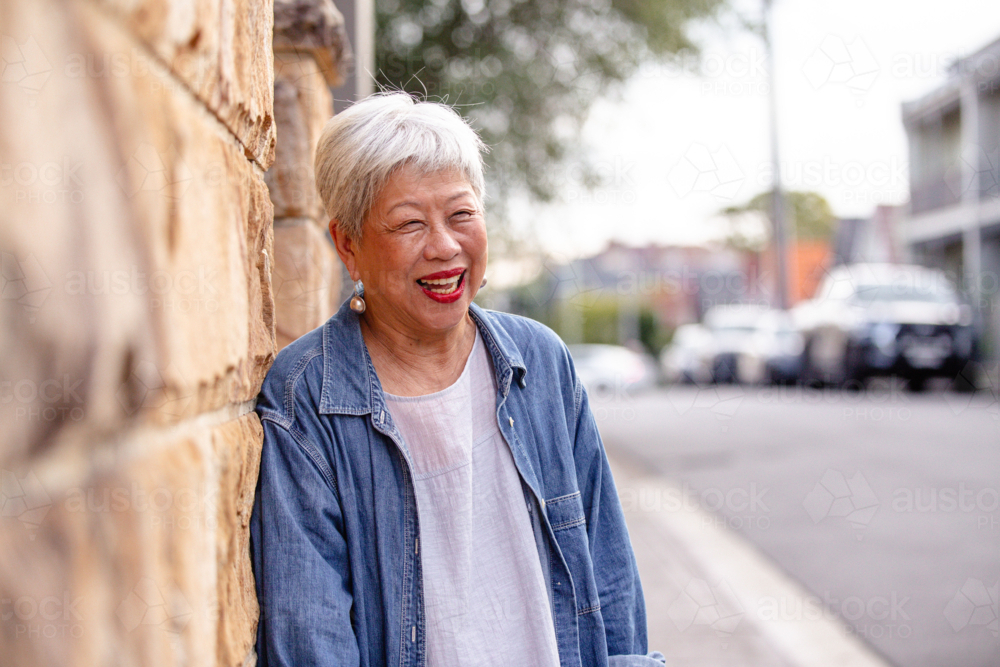 Senior Asian woman laughing while leaning against the wall at the roadside. - Australian Stock Image