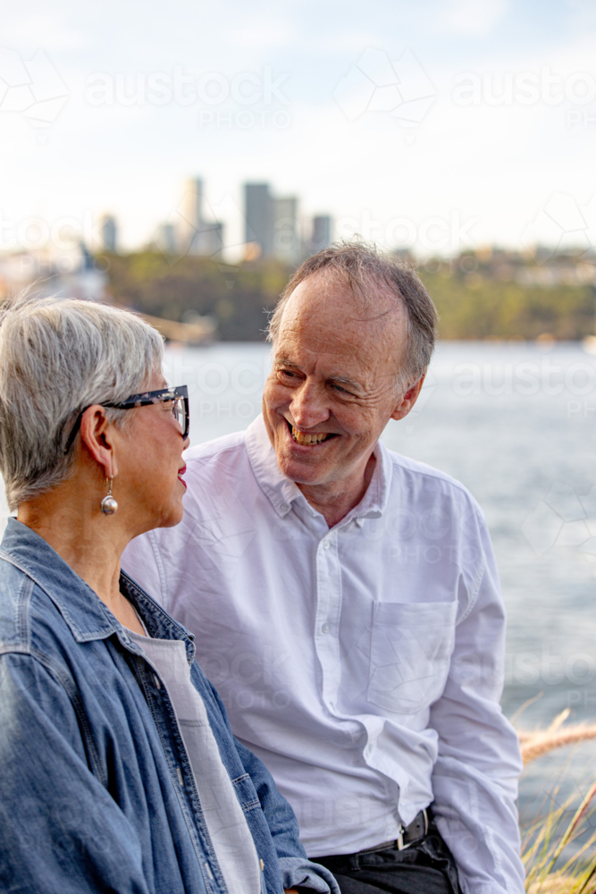 Senior man looking at Asian woman while sitting outdoors near the water - Australian Stock Image