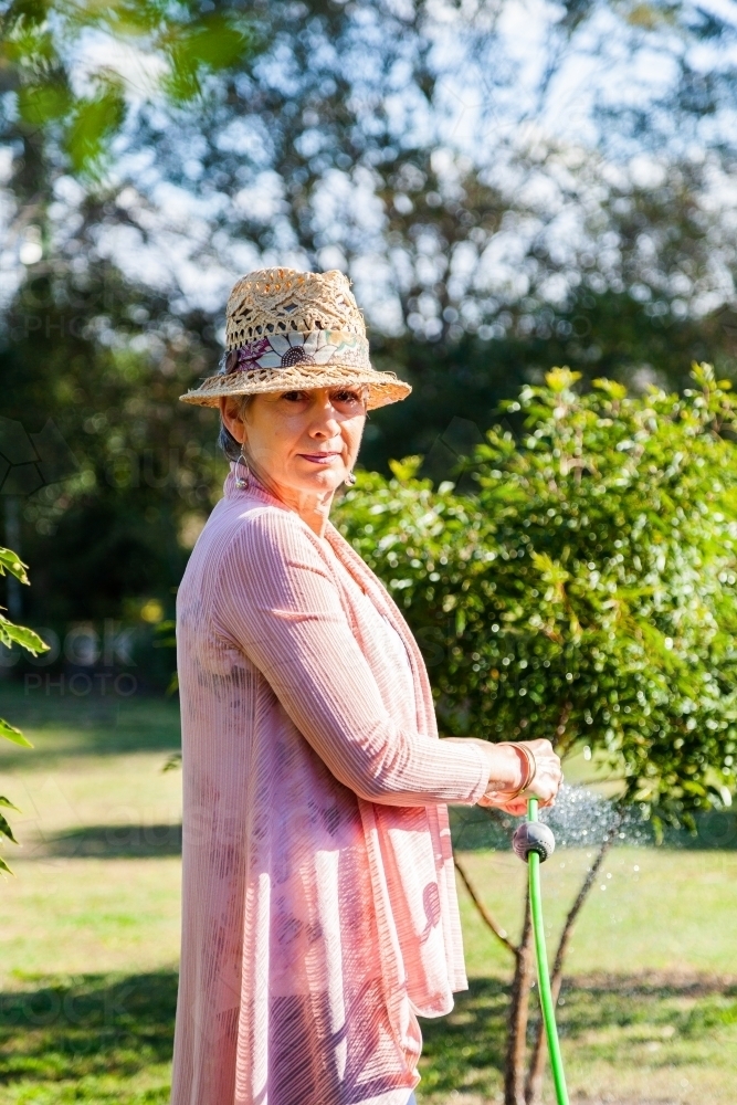 Senior woman watering plants in the garden with green hose - Australian Stock Image