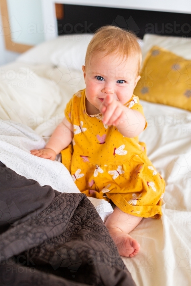 Seven month old baby sitting on parents bed pointing and smiling - Australian Stock Image