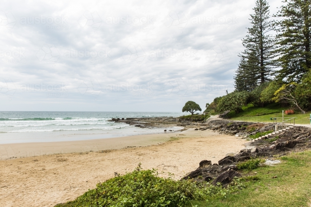 Shore of ocean beach with cloudy skies and grass and trees - Australian Stock Image