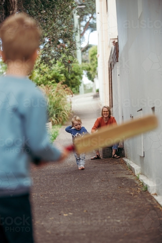 Shot from behind of young boy playing cricket in the street with his family - Australian Stock Image