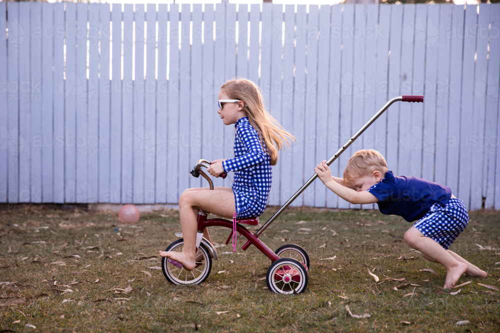 siblings playing on a trike in the back yard with a girl riding and a boy pushing - Australian Stock Image