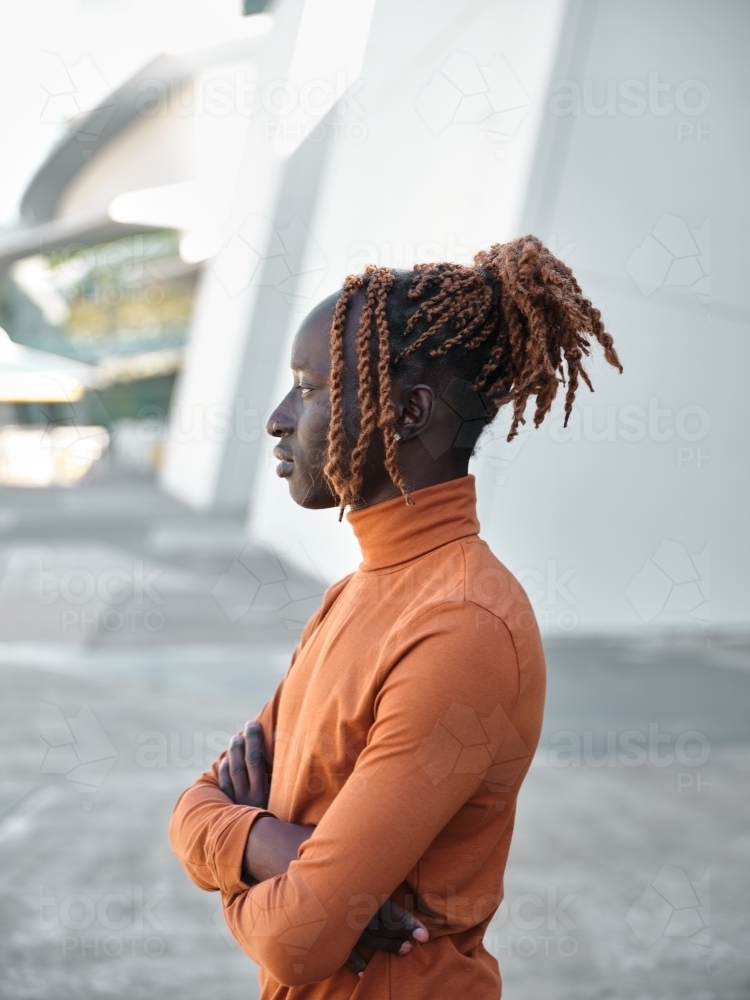 Side profile of South Sudanese man wearing orange turtleneck with arms folded - Australian Stock Image