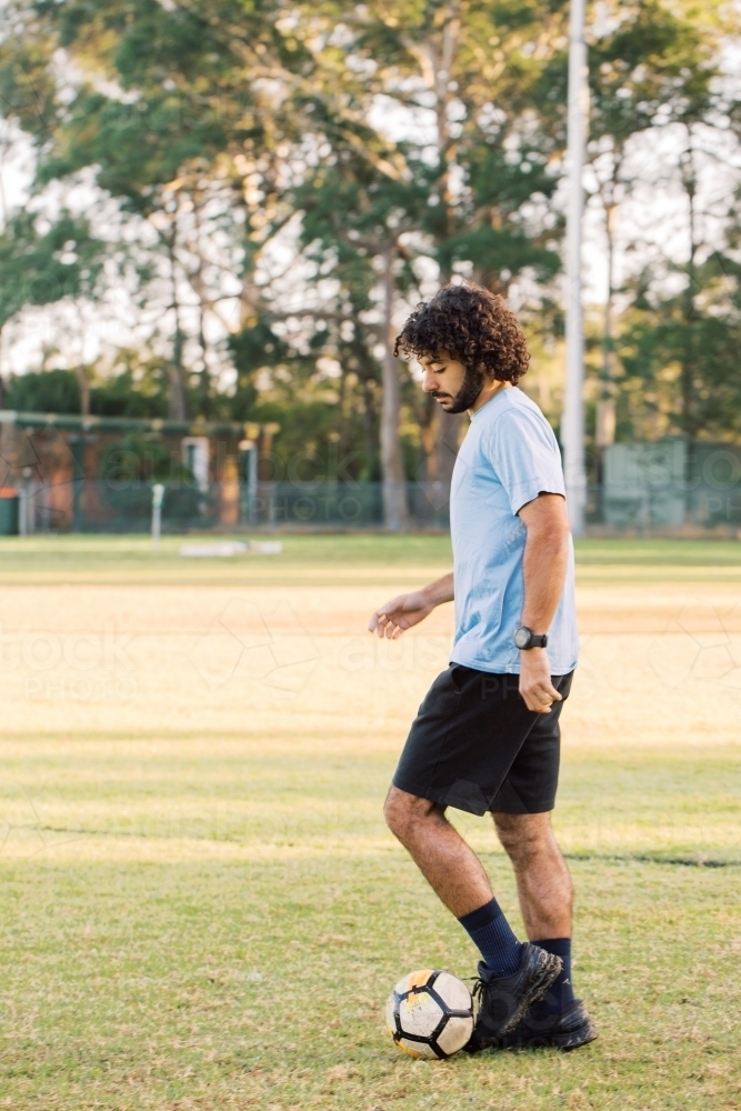 Side view shot of a man with curly hair wearing sports clothes playing a soccer ball on the field - Australian Stock Image