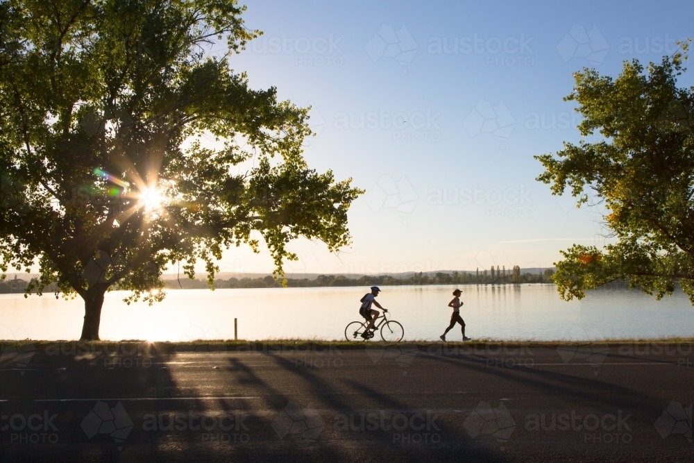 Silhouette of a woman running and a man cycling beside Lake Burley Griffin - Australian Stock Image