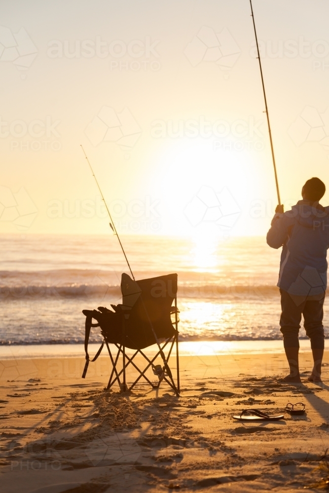 Silhouette of man and camp chair on seashore fishing at sunrise - Australian Stock Image