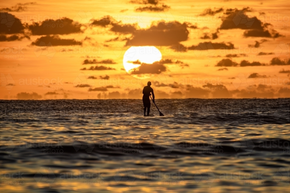 Silhouette of paddler with the large sun rising behind him on the ocean water - Australian Stock Image