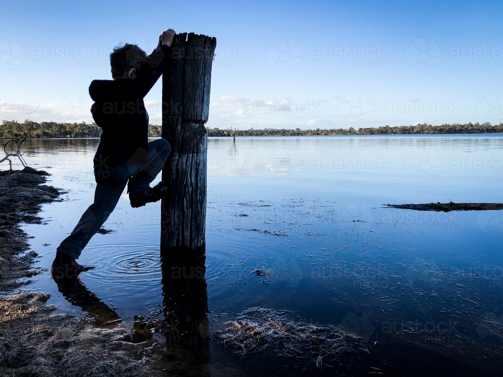 Silhouette of young boy attempting to climb pole immersed in glassy lake water in winter clothing - Australian Stock Image