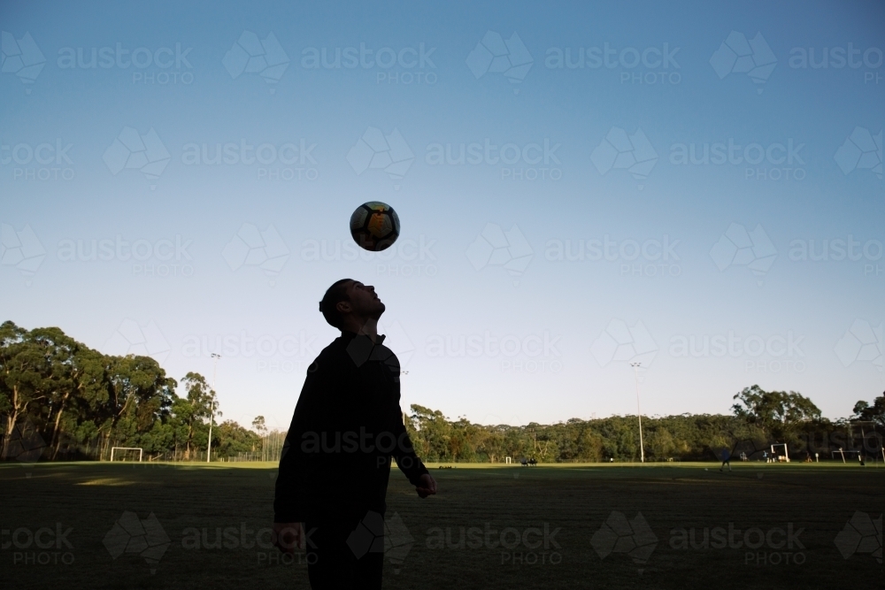 Silhouette shot of a man, head bouncing a ball soccer ball in the field with blue skies - Australian Stock Image