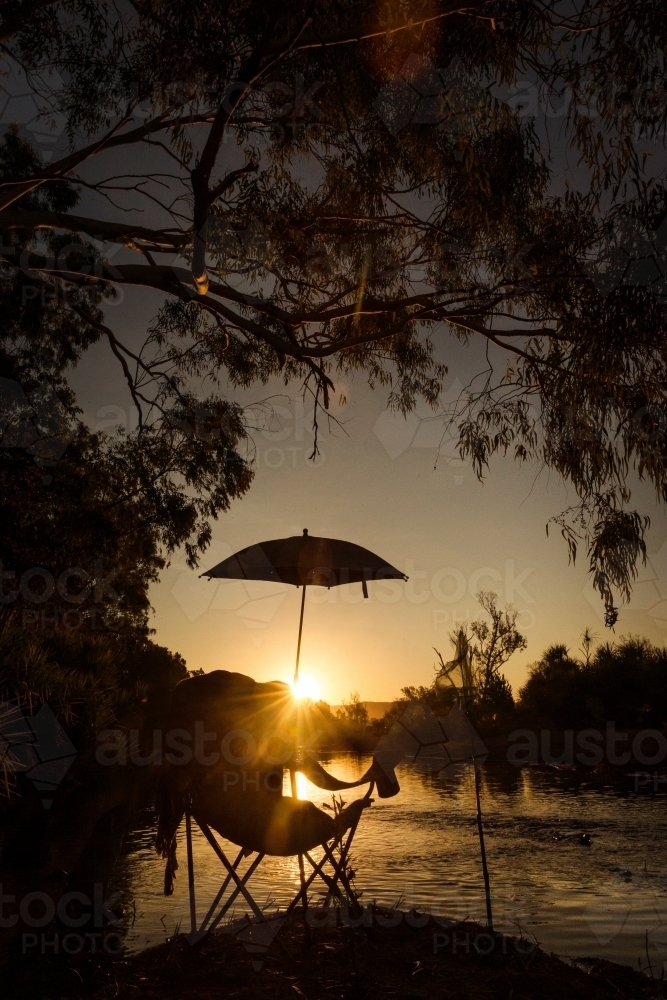 silhouette shot of lake with foldable chair and umbrella under a tree during a sunset - Australian Stock Image