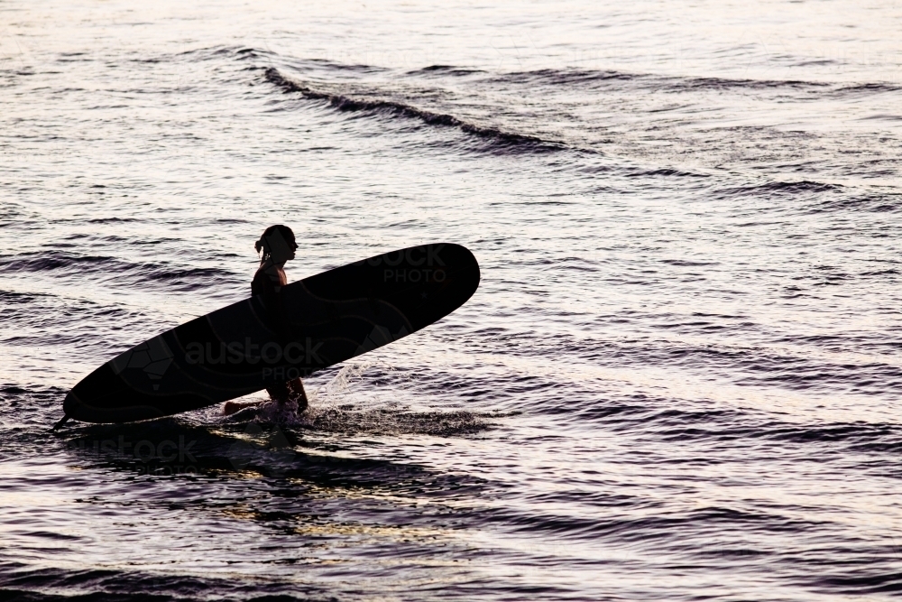 Silhouetted female surfer holding a longboard entering the water at dusk, Byron Bay. - Australian Stock Image