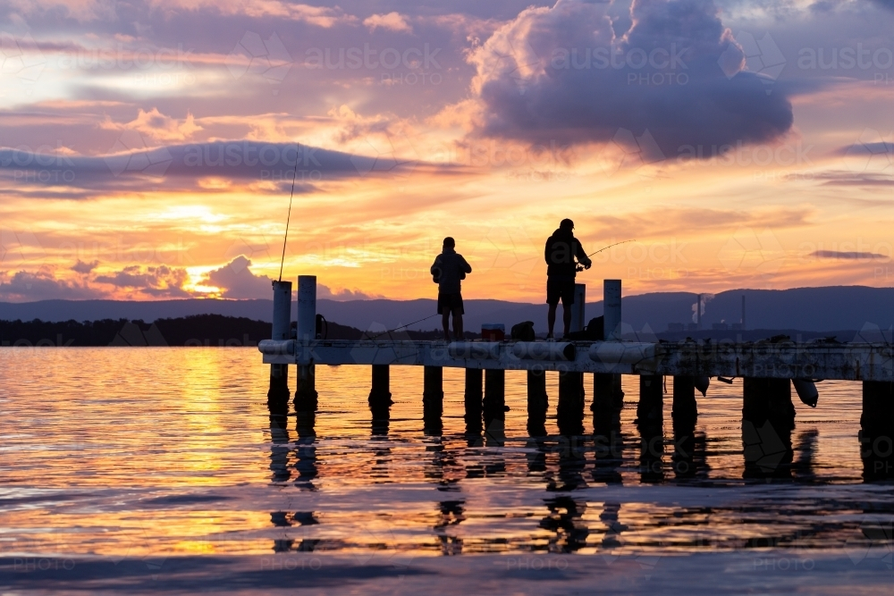 Silhouetted fishermen fishing on jetty at sunset - Australian Stock Image