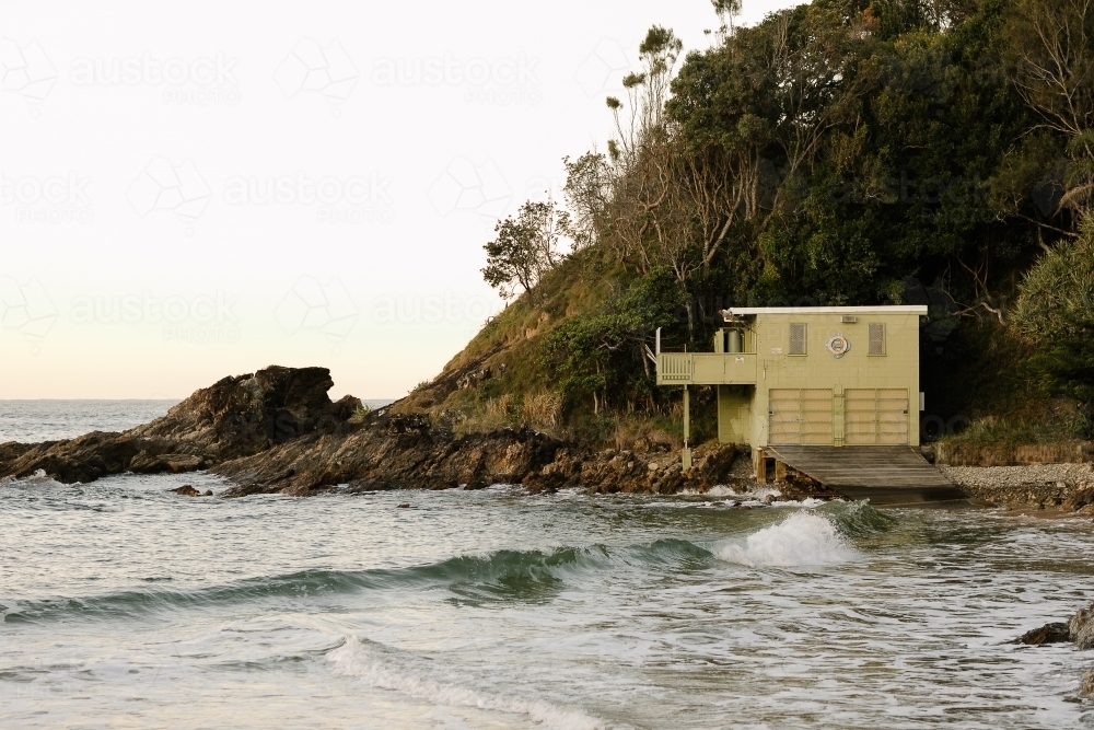 Small green boat shed on the beach with small waves and a hill behind it - Australian Stock Image