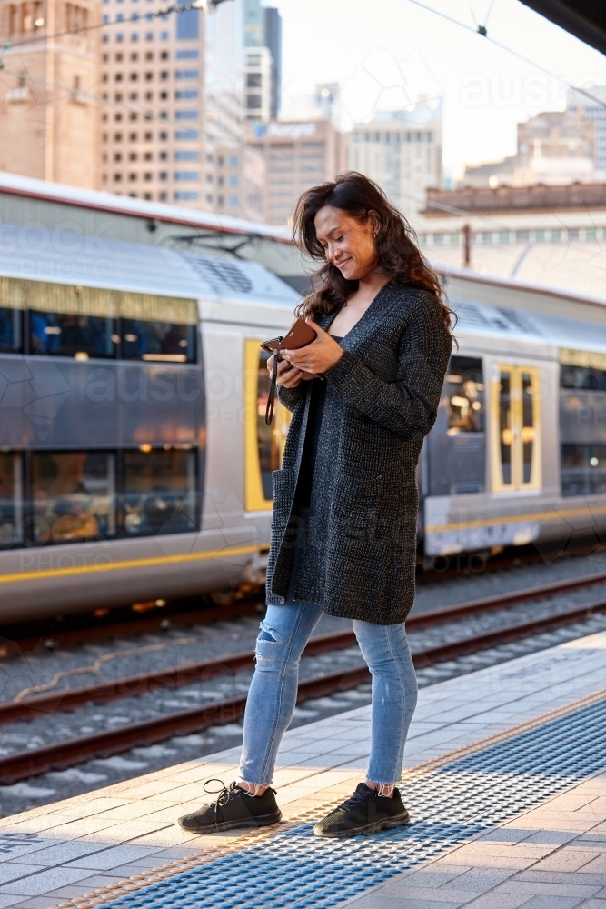 Smiling Asian woman waiting at train station with mobile phone - Australian Stock Image