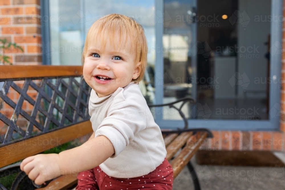 Smiling baby girl sitting on park bench outside on back porch of home - Australian Stock Image