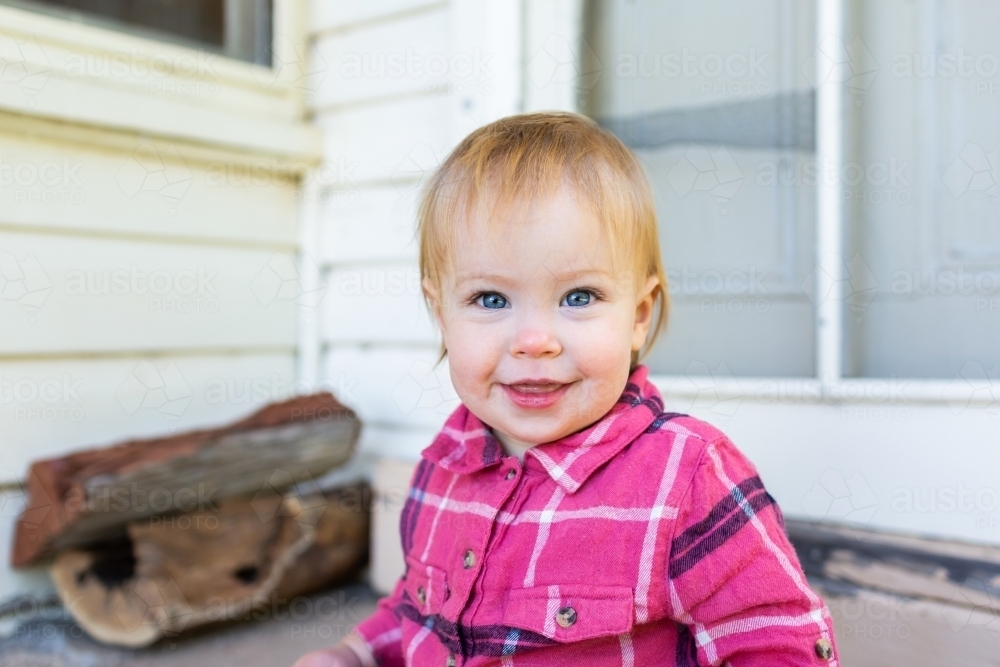 Smiling country girl with blue eyes and blonde hair sitting on back doorstep of country farm house - Australian Stock Image