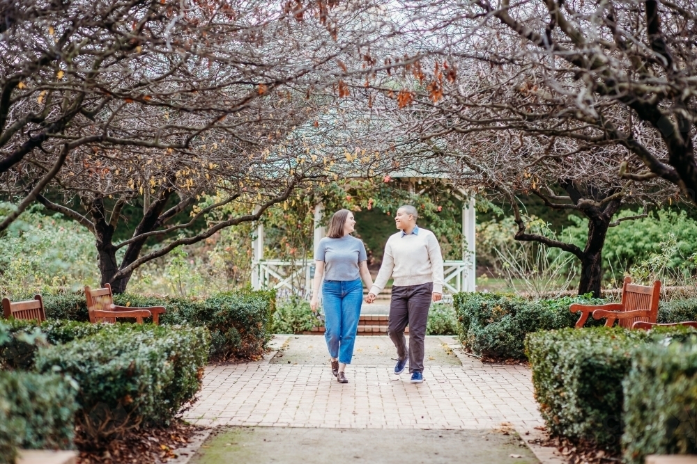 smiling lgbt  couple holding hands with dead trees, shrubs and wooden benches on the side - Australian Stock Image