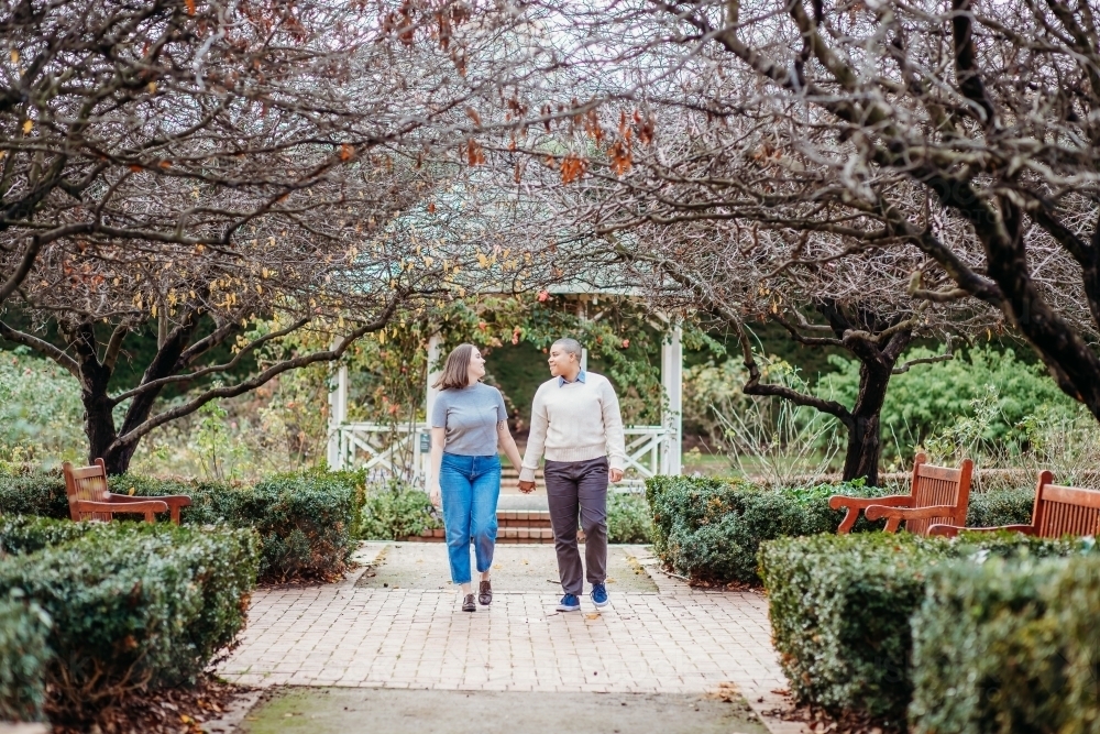 smiling lgbt  couple holding hands with dead trees, shrubs and wooden benches on the side - Australian Stock Image