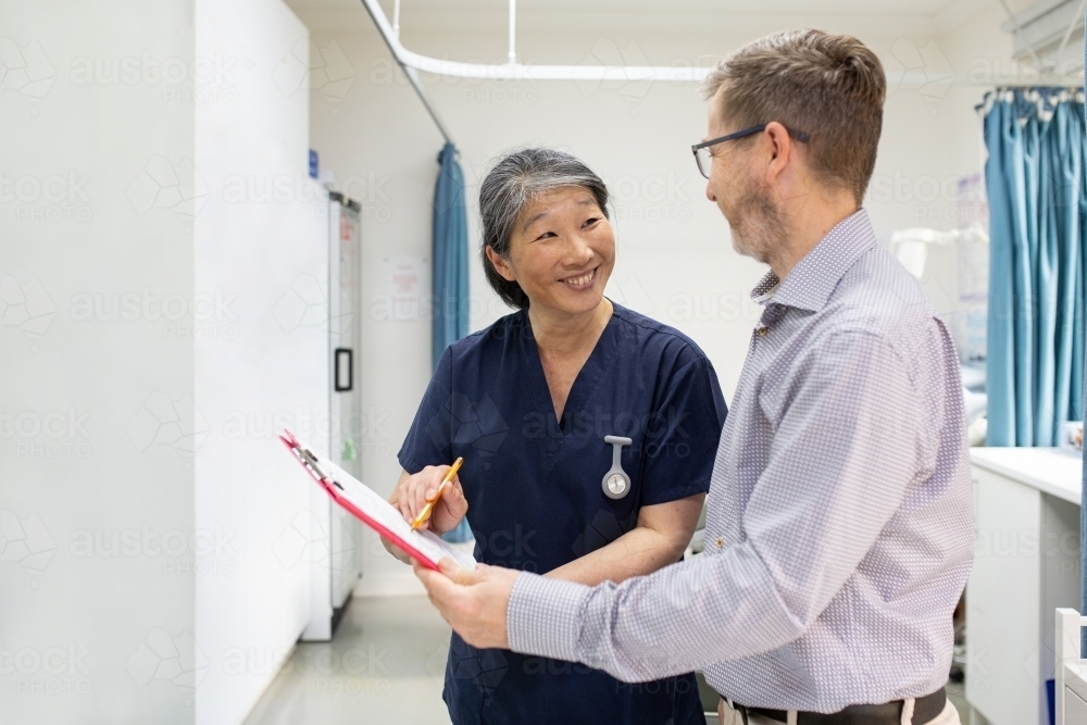 smiling middle aged woman wearing blue scrubs holding a file while talking to a man in a clinic ward - Australian Stock Image