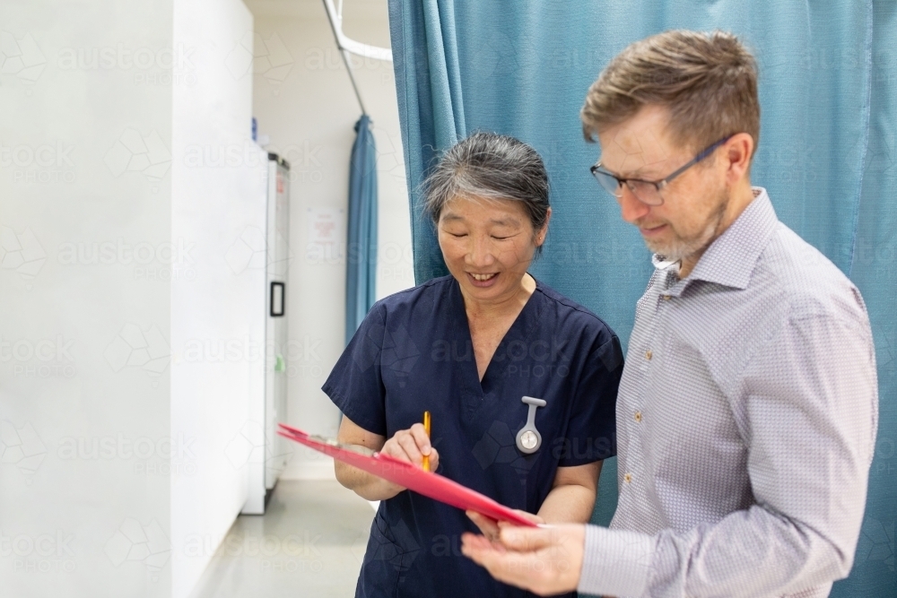 smiling middle aged woman wearing blue scrubs holding a file while talking to a man in a clinic ward - Australian Stock Image