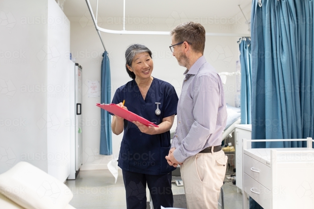 smiling middle aged woman wearing blue scrubs holding a file while talking to a man in a clinic ward - Australian Stock Image