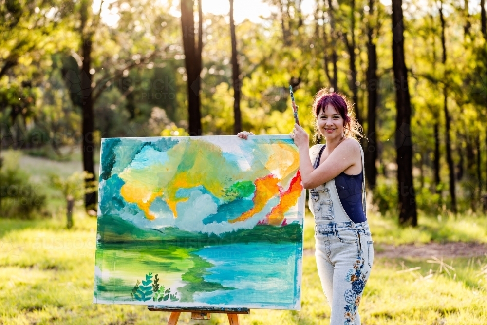 Smiling portrait of happy young woman artist in forest clearing with beginning of painting - Australian Stock Image