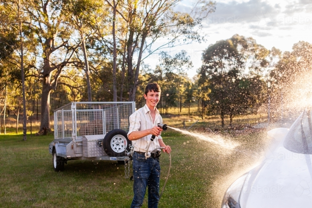 Smiling teen boy washing car spraying vehicle with water - Australian Stock Image