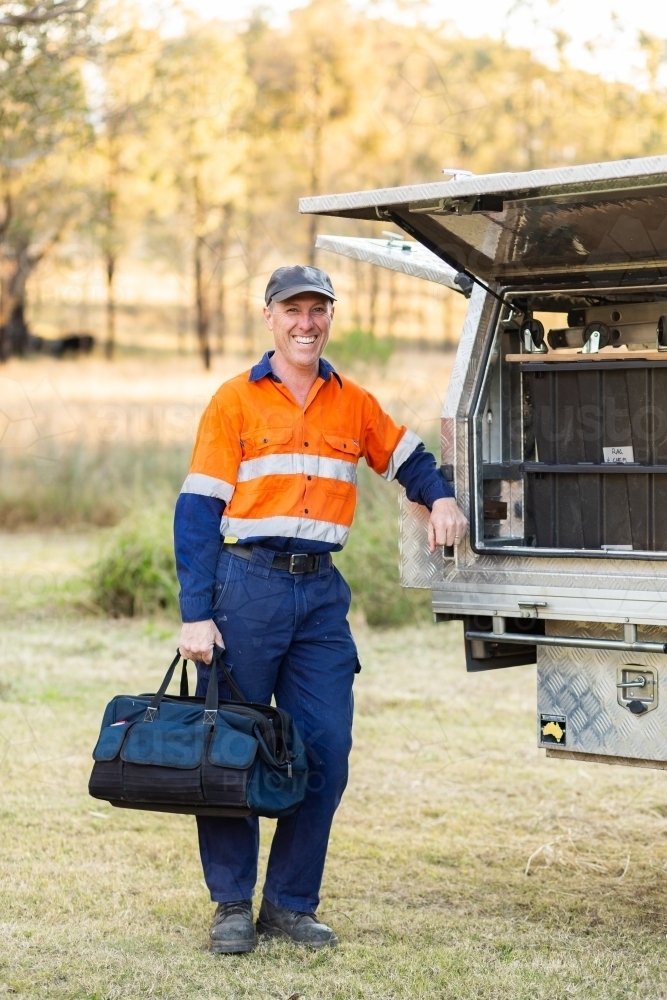 Smiling tradie leaning on work ute with bag of tools - Australian Stock Image