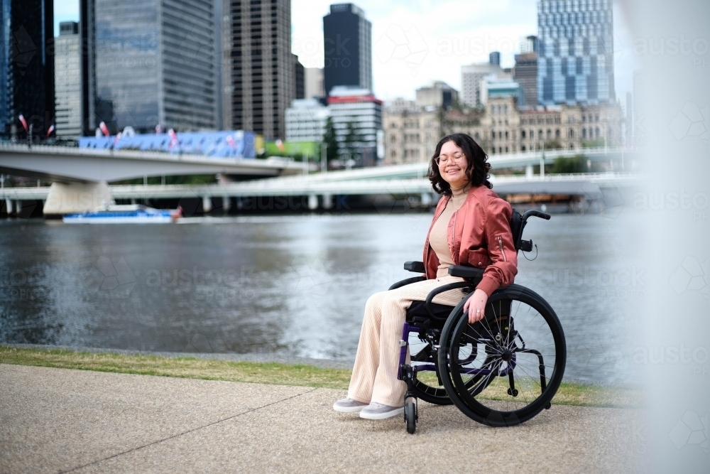 Smiling woman with her eyes closed  in wheelchair beside river - Australian Stock Image