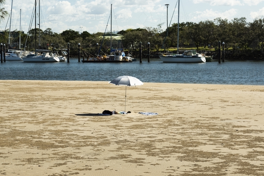 Solitary solitary beach umbrella in beach scene with boats moored in background at riverside - Australian Stock Image