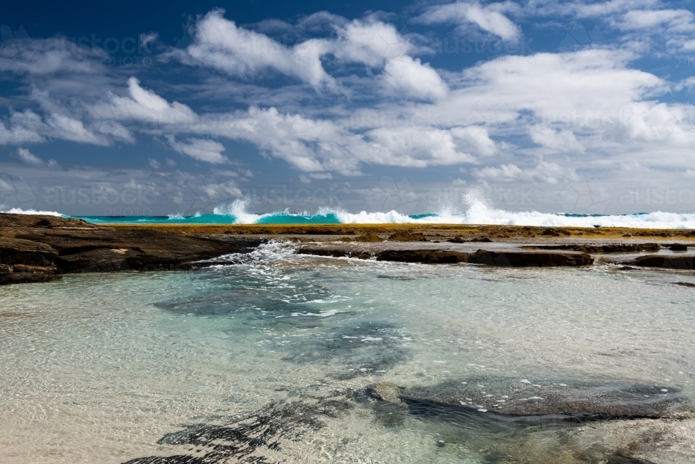 Spectacular low view of water flowing into rock pool with waves behind and dramatic blue cloudy sky - Australian Stock Image