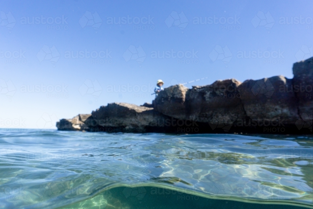 Split shot of out of focus boy wearing hat playing with fishing rod on rocks near water - Australian Stock Image