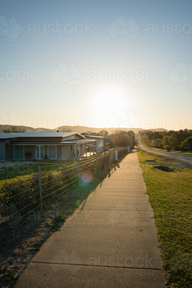 Suburban street glows in warm sunset, casting long shadows on the empty footpath - Australian Stock Image