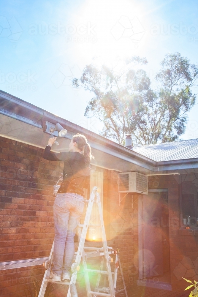 Sun flare over ladie tradie on a ladder painting the exterior of a home - Australian Stock Image